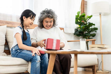 Asian young little girl give gift to grandmother at home. They sit on sofa together. Old smile happy woman open box with granddaughter. Two generation have happiness moment. Family lifestyle concept.の写真素材