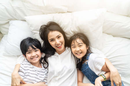 Asian cute family mother, son and daughter lying on bed with happiness and smile in bedroom. Little young boy and girl hug mom smiling enjoy playing together on white bed sheet,  love and worm touch.の写真素材