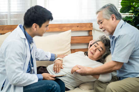Asian senior old woman with bad condition feeling sick lying on sofa having medical check up by doctor visiting at home. Elder man husband taking care of wife closely and worry about unwell illness.の写真素材
