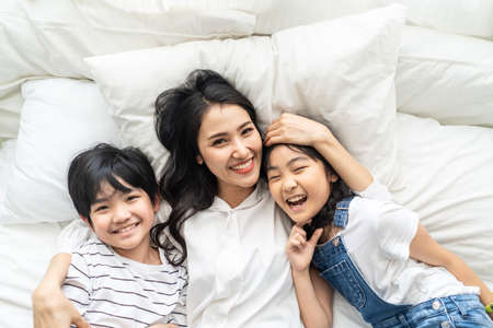 Asian cute family mother, son and daughter lying on bed with happiness and smile in bedroom. Little young boy and girl hug mom smiling enjoy playing together on white bed sheet,  love and worm touch.の写真素材