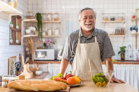 Senior Asian elder man happy living in home kitchen. Grandfather cooking salad dish standing at counter with happiness and smile enjoy retirement life. Older people activity in family at home.の写真素材