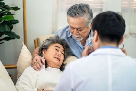 Asian senior old woman with bad condition feeling sick lying on sofa having medical check up by doctor visiting at home. Elder man husband taking care of wife closely and worry about unwell illness.の写真素材