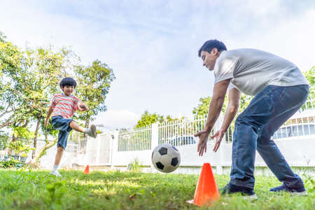 Asian young family playing football in garden playground at home together. Son shooting a ball to father who are goalkeeper on green grass. Dad stand hand to catching ball at backyard. Family activityの写真素材