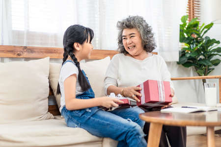 Asian young little girl give gift to grandmother at home. They sit on sofa together. Old smile happy woman open box with granddaughter. Two generation have happiness moment. Family lifestyle concept.の写真素材