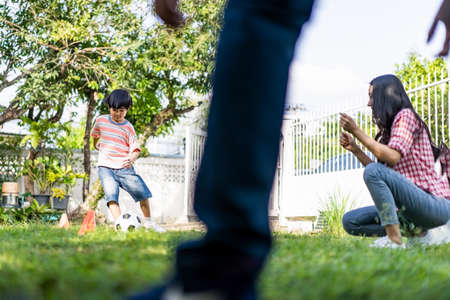 Asian young family playing football in garden playground at home together. Son running to kick ball pass orange cone to dad who are goalkeeper on green grass while mother cheer boy up. Happy activityの写真素材