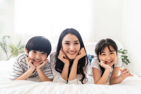 Asian family mother, son and daughter lying on white bed with happiness and smile in bedroom. Mom, boy and girl playing together feels fun. People looking camera. House property mortgage advertisementの写真素材