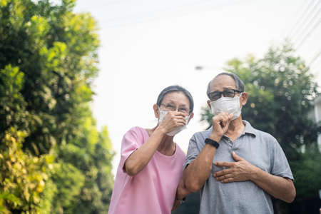 Asian elder senior couple wearing mask for prevent dusk pm 2.5 bad air pollution. Old woman and man holding mask and having a cough due to bad smell, allergic sickness symptom stand in front of home.の写真素材