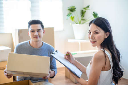 Asian family moving house, young cute woman checking things on paper on clipboard while man carrying boxes. Wife and husband looking at camera and smile with happy face. Home moving and delivery.の写真素材