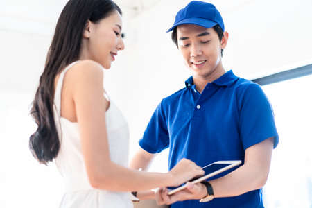 Asian postman deliver item product package to young girl at home. People happy to get post parcel fast, comfortable using express transportation delivery service. Woman sign name on deliveryman tabletの写真素材