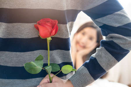 Asian young man making surpirse by hiding red rose flower behind before giving to pretty girlfriend. Woman waiting present with smile. Focus on beautiful flower. Love couple relationship concept.の写真素材