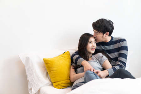Lovely Asian couple sitting on bed in bedroom together. Boy holding girl head softly and be hand in hand or pinky swear to love girl. Man kissing woman on smile young lady cheek feeling shy & happy.の写真素材