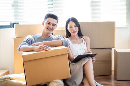 Portrait of Asian family moving house, young cute woman and handsome man holding box sitting on floor. Wife and husband looking at camera and smile with happy face. Home moving and delivery concept.の写真素材