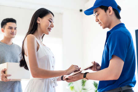 Asian postman delivered object package to young couple at home. People happy to get parcel post very fast, good care of fragile inside by Express Transportation. Woman sign name on tablet deliveryman.の写真素材