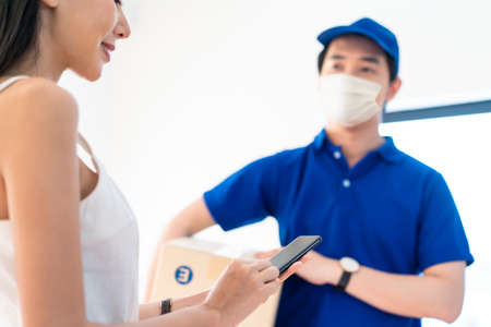 Asian postman or deliveryman carry small box deliver to young woman customer in front of door at home. Man wearing mask prevent covid or coranavirus affection outbreak. Social distancing work concept.の写真素材