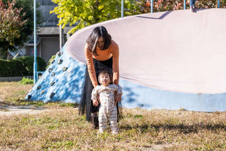 Asian mother holding little cute toddler in park teaching her child how to walk. Kid got helping from mom to stand and take first step forward. Baby and child education development concept.の写真素材