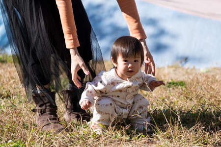 Asian mother holding little cute toddler in park teaching her child how to walk. Kid got helping...