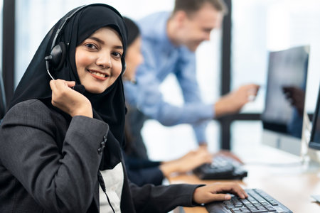 Call center Asian Muslim woman working in office with colleagues. The girl holding headset microphone and smile to camera. Her co-workersÂ  working behind as background. Customer service concept.の写真素材