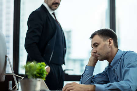 Caucasian boring male worker or employee sitting on desk typing on laptop feeling stress and bored. Man working under pressure by their boss feeling angry standing behind forcing them complete job.の写真素材