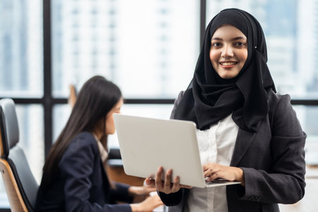 Young Asian Muslim girl holding laptop standing in working office with smile face. Colleague sitting typing on computer behind. Workplace is in downtown area with high rise building view in backgroundの写真素材
