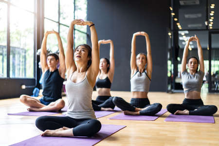 Group of Multi ethics people learning Yoga class in fitness club. Female Asian instructor leading Meditate sitting stretch hand and arms pose, students follow behind. Yoga for health, wellness conceptの写真素材