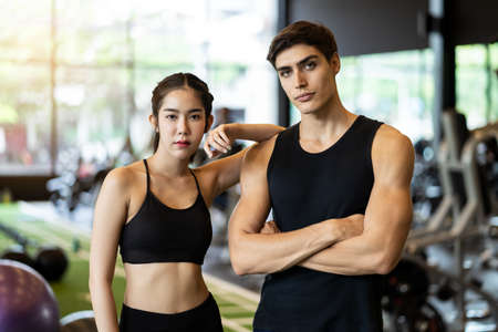 Portrait of young handsome fit Caucasian man and beauty Asian woman standing showing muscle body in gym or fitness club with exercise equipment in background. Working out for health, well being.の写真素材