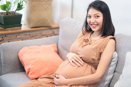 Young happy pretty Asian pregnant woman sitting and looking at camera with smile holding and touching her tummy thinking about future of the her baby. Pregnancy woman health concept.の写真素材
