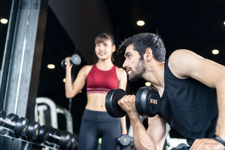 Young Caucasian handsome and pretty Asian girl are lifting the weights in gym or fitness club. They both have very strong and good body shape. Seen side of the man is looking straight in the mirror.の写真素材