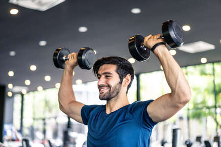 Young fit Caucasian handsome man lifting weights dumbbell both hands in gym or fitness club. Male athlete exercise on shoulder and arm muscle having good body shape sitting on bench, healthy concept.の写真素材
