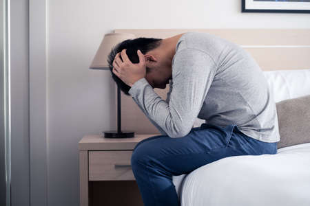Stress Asian young man sitting alone on bed. He crying with tear and put both hands on head. He is...
