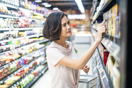 Young Asian beautiful woman holding grocery basket walking in supermarket. She is looking and...