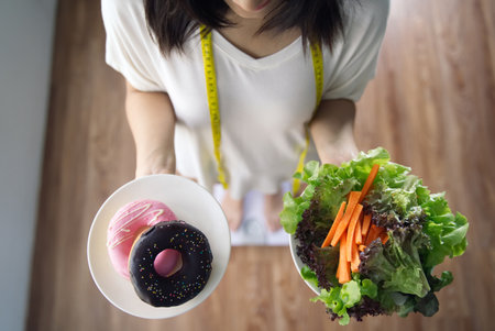 Top view of young Asian girl holding dishes of vegetable salad and donuts standing on weight measurement scale with measure tape on neck. Decision making  for her health. Diet for geed health concept.の写真素材