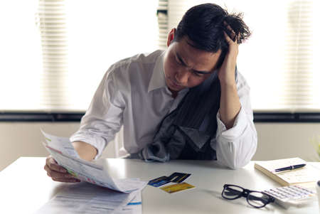 Stressed young Asian man with business office wear holding bills and another hand on head, thinking about  finding money to pay credit card debt and all bills. Financial problem concept.の写真素材