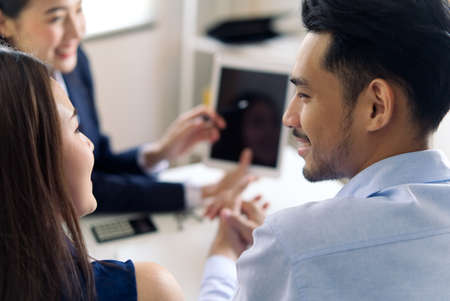 Investment female adviser giving a presentation to an Asian friendly smiling young couple seated at her desk in the office. The couple looking each other with smile after listening agent explaining.の写真素材