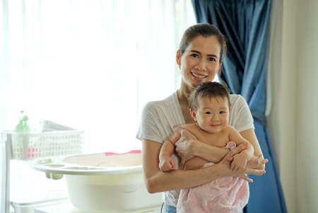 Asian beautiful mother holding little cute baby after taking a bath and dry her kid with towel standing in the room. Bathtub and bath shampoo are put in the background. Clean and healthy concept.の写真素材