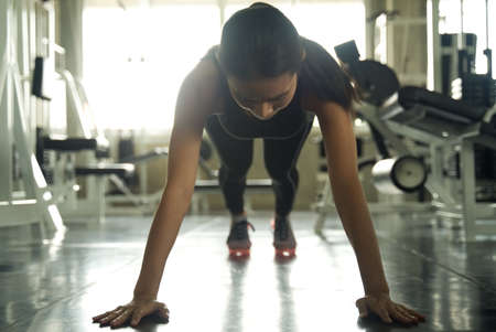Young Asian beautiful girl working out in the gym. She is doing a plank for abdominal muscles training. Seen from her front with training equipment in background. Health and fitness concept.の写真素材