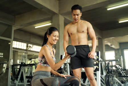 Young Asian beautiful girl working out in the gym. She is sitting and lifting the dumbbell and having her male friend coaching how to lift it correctly behind. Health and fitness concept.の写真素材