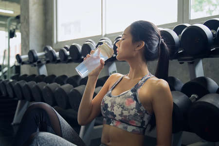 Beauty Asian young girl drinking water in fitness after she had some work out. She sitting in front of dumbbells rack and holding the bottle of water. Seen from the side. Gym and fitness concept.の写真素材