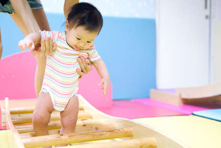 Asian baby taking first steps walk forward on the soft mat. Happy little baby learning to walk with...