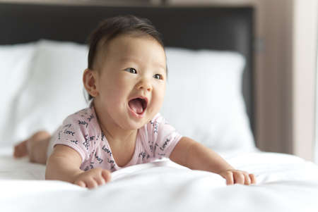 Young cute Asian baby crawling on bed in bedroom. The baby is smiling and looking straight at mother...
