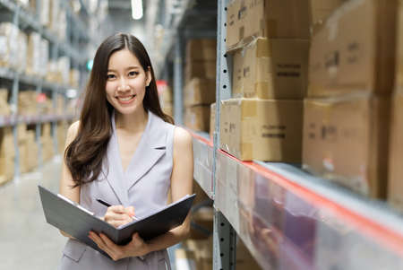 Smart smiling Asian woman working in storage warehouse. She is standing and holding document folder and writing some note. She is feeling happy to work. Smart working office girl concept.の写真素材