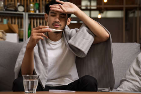Asian young man with blanket getting a cold or flu sitting on sofa at home. He is having a fewer checked by thermometer and put one hand on head. Illness and sickness concept.の写真素材