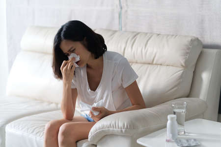 Unwell condition of Asian women sitting on sofa. She has a Runny nose so she blowing the nose and...