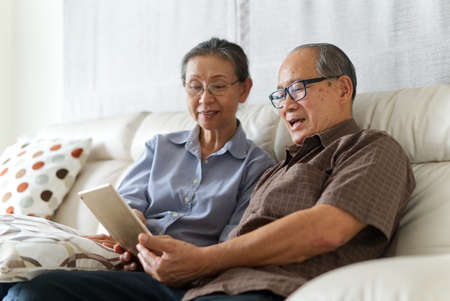 Senior couple sitting on sofa in home playing tablet and relaxing together. They are smiling and enjoy to spend their time together with happiness. Happy retirement life concept.の写真素材
