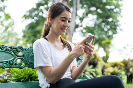 Asian woman enjoy communicate chatting on mobile phone in public park. Young active beautiful girl in sportswear resting, take a break after exercise by jogging workout outdoors in evening at garden.の写真素材