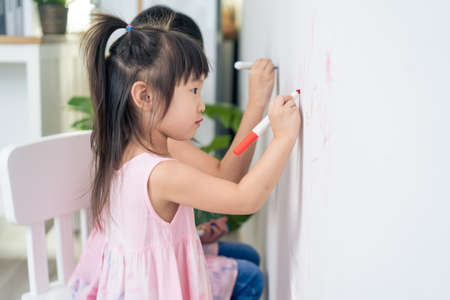 Asian young sibling kid girl enjoy paint on white wall in living room. Little adorable children having fun drawing and coloring art picture with hapiness enjoy creativity activity on holiday at home.の写真素材