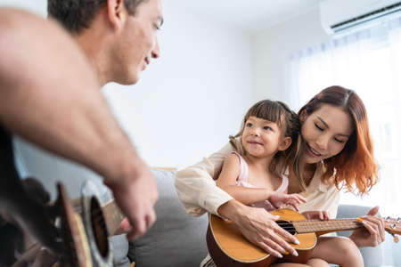 Asian young couple playing guitar with young baby kid together at home. Attractive beautiful parents sit on sofa, spend leisure time with little child daughter singing a song in living room in house.の写真素材