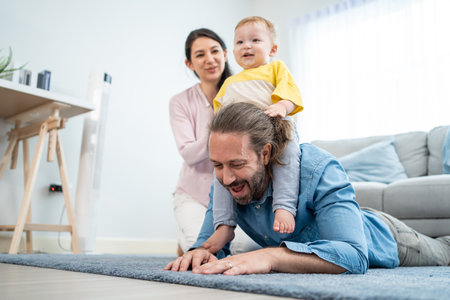 Caucasian happy loving parent play with baby toddler in living room. Attractive couple father carry young little infant son child on neck with mother on floor. Activity relationship at home in house.の写真素材