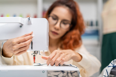 Asian beautiful tailor woman working on clothes in tailoring atelier. Attractive young female fashion designer pushes fabric material through a sewing machine making new handmade suit in workshop roomの写真素材
