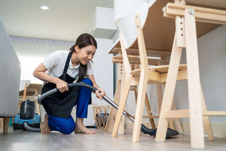 Asian cleaning service woman worker cleaning in living room at home. Beautiful young girl housekeeper cleaner wear apron and vacuuming messy dirty floor for housekeeping housework and chores in house.の写真素材