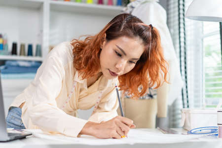 Asian beautiful tailor woman working on clothes in tailoring atelier. Attractive female fashion...
